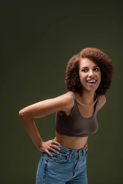 Femme afro-américaine positive en haut et jeans regardant loin isolé sur vert foncé — Photo de stock