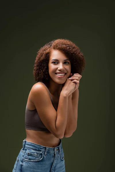 Feliz y rizado modelo afroamericano en la parte superior y jeans mirando hacia otro lado aislado en verde oscuro - foto de stock