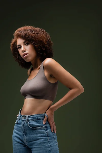 Ricci donna afroamericana in alto e jeans che tengono la mano sull'anca e guardando la fotocamera isolata sul verde scuro — Foto stock