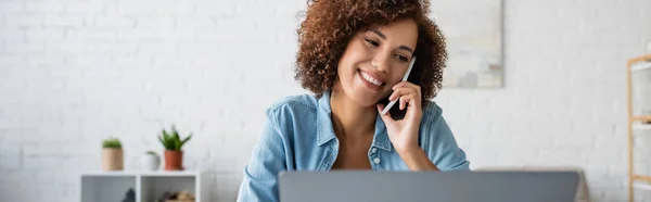 Mujer afroamericana positiva hablando en el teléfono móvil cerca de la computadora portátil borrosa en casa, pancarta - foto de stock