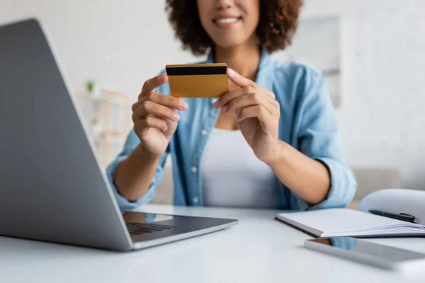Vista recortada de mujer afroamericana borrosa sosteniendo tarjeta de crédito cerca de dispositivos - foto de stock