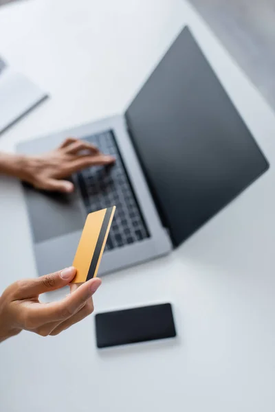 Vue recadrée d'une femme afro-américaine tenant une carte de crédit et utilisant un ordinateur portable avec écran blanc à la maison — Photo de stock