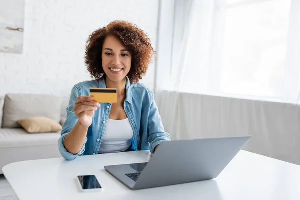Mujer afroamericana positiva sosteniendo tarjeta de crédito cerca de dispositivos en casa - foto de stock