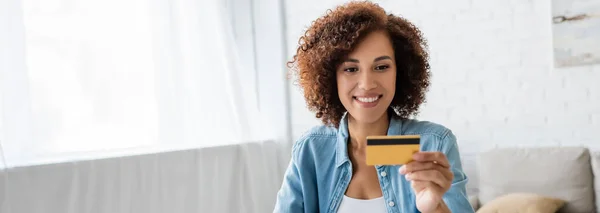 Улыбающаяся африканская американка смотрит на кредитку в гостиной, баннер — стоковое фото