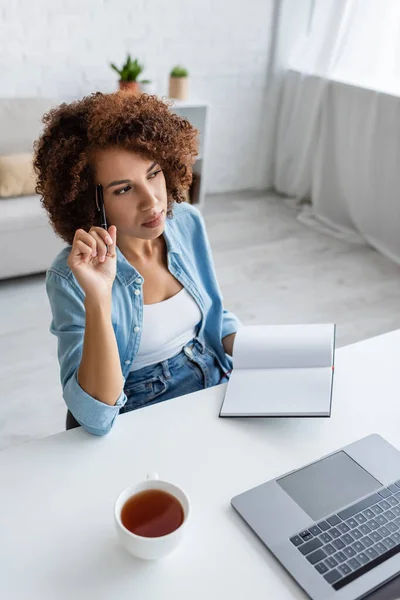 Pensativo freelancer afroamericano sosteniendo portátil cerca de la computadora portátil y una taza de té en casa - foto de stock