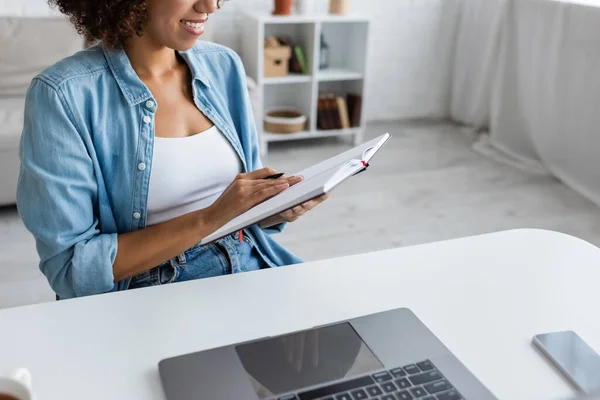 Vista recortada del alegre freelancer afroamericano sosteniendo un cuaderno cerca de dispositivos con pantalla en blanco en casa - foto de stock