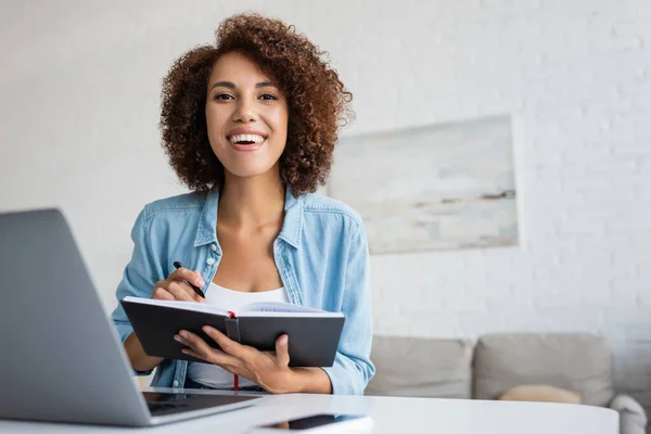 Happy african american freelancer looking at camera and holding notebook at home — Stock Photo