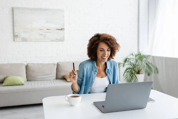 Smiling african american freelancer holding pen near devices and cup of tea at home — Stock Photo