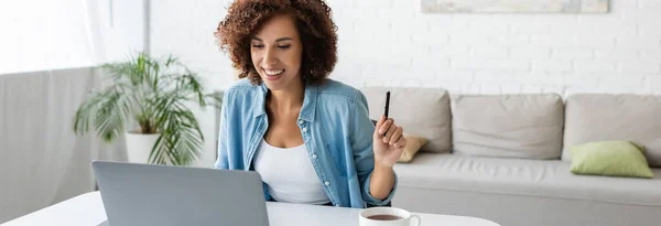 Donna afroamericana allegra che tiene la penna e guarda il computer portatile vicino al tè a casa, banner — Foto stock