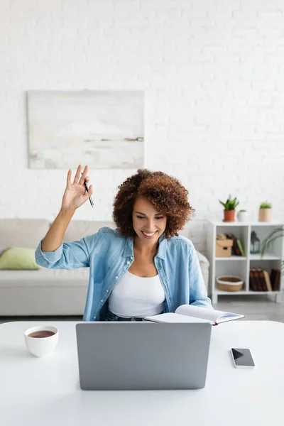 Sonriente freelancer afroamericano con portátil saludando la mano durante la videollamada en el ordenador portátil en casa - foto de stock