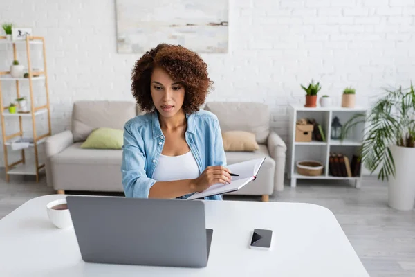 Curly african american woman holding notebook and using laptop near cup of tea at home — Stock Photo