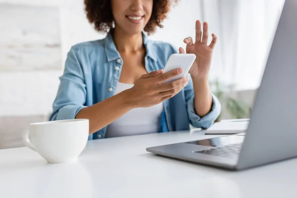 Cropped view of african american freelancer using smartphone near laptop and cup at home — Stock Photo