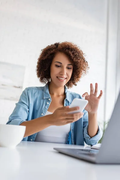 Smiling african american freelancer using smartphone near laptop and blurred cup on table — Stock Photo