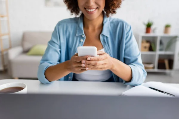 Vista cortada de freelancer afro-americano alegre usando smartphone perto de laptop e chá em casa — Fotografia de Stock