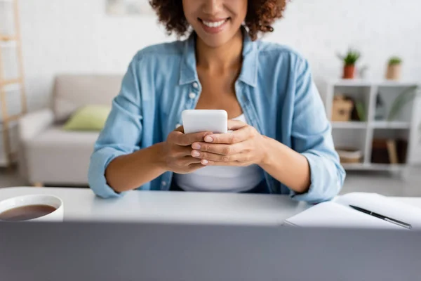 Ausgeschnittene Ansicht der fröhlichen afrikanisch-amerikanischen Freelancer mit Smartphone in der Nähe von Laptop und Tee zu Hause — Stockfoto