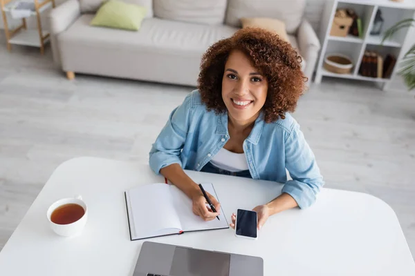 Cheerful african american woman holding smartphone and writing on notebook near laptop at home — Stock Photo