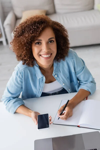 High angle view of african american freelancer holding smartphone near notebook at home — Stock Photo