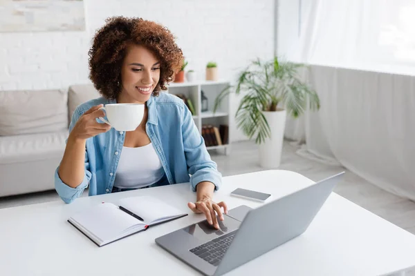 Positivo donna afro-americana in possesso di tazza e utilizzando computer portatile vicino notebook sul tavolo a casa — Foto stock