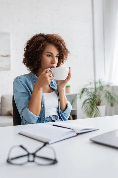 Curly african american woman drinking coffee and looking at laptop on desk — Stock Photo