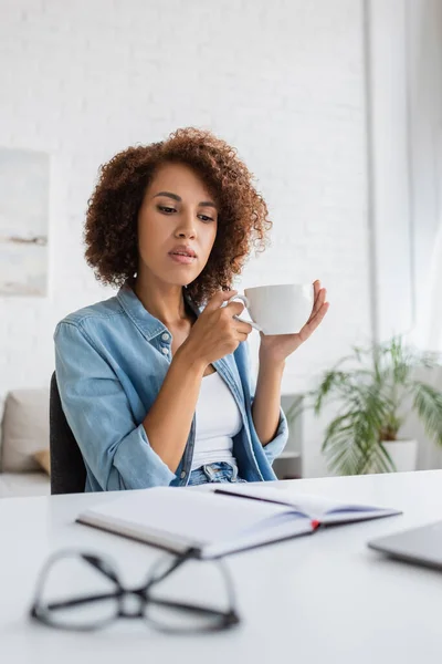 Pensivo Africano americano mulher segurando xícara de café e olhando para notebook na mesa — Fotografia de Stock