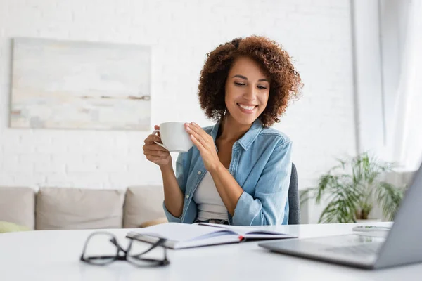 Fröhliche afrikanisch-amerikanische Frau hält eine Tasse Kaffee in der Hand und blickt auf ein Notizbuch neben dem Laptop auf dem Schreibtisch — Stockfoto