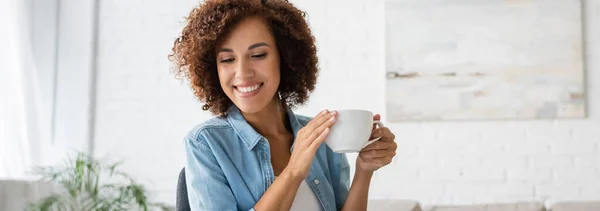 Fröhliche afrikanisch-amerikanische Frau, die eine Tasse Kaffee in der Hand hält und zu Hause wegschaut, Banner — Stockfoto