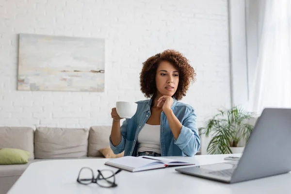 Nachdenkliche afrikanisch-amerikanische Frau hält Tasse Kaffee in der Hand und blickt auf Laptop auf Schreibtisch — Stockfoto