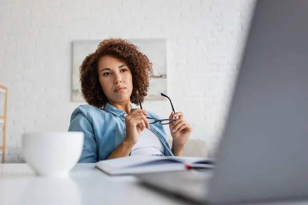 Curly african american woman holding glasses and looking at blurred laptop — Stock Photo