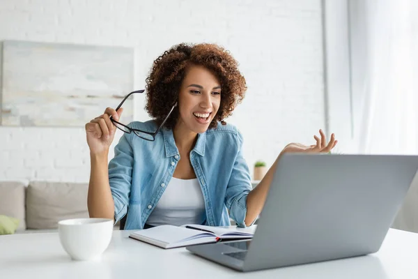 Mujer afroamericana feliz sosteniendo gafas y mirando a la computadora portátil en la mesa - foto de stock