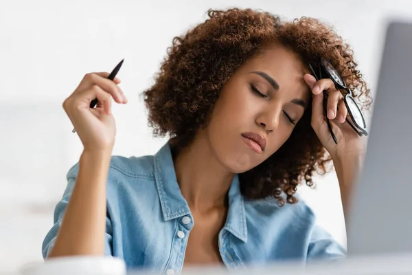 Tired african american woman with closed eyes holding glasses and pen — Stock Photo