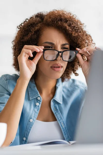 Focused african american woman wearing glasses and looking at blurred laptop — Stock Photo