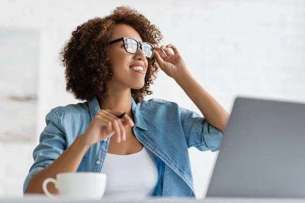 Mujer afroamericana feliz con gafas y mirando hacia otro lado cerca de la computadora portátil - foto de stock