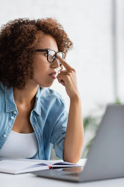 Encaracolado afro-americano mulher ajustando óculos e olhando para longe perto de laptop borrado — Stock Photo