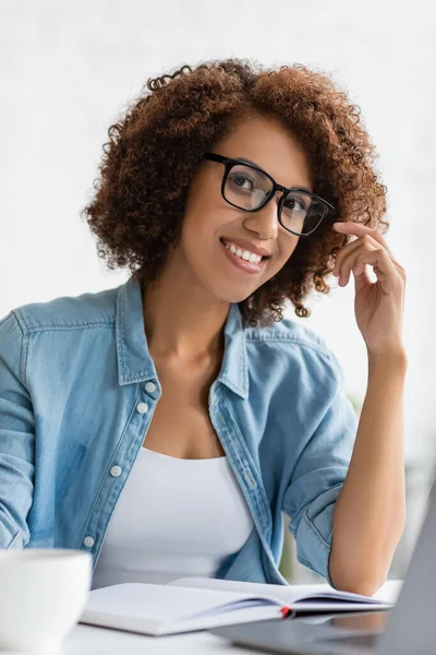 Счастливая африканская американка в очках, смотрящая в камеру, сидя за столом — стоковое фото