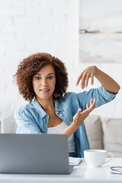 Curly african american woman gesturing during video call on laptop near cup on desk — Stock Photo
