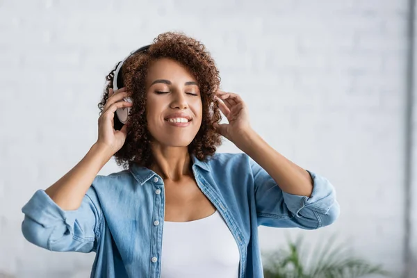 Mujer afroamericana rizada sonriendo mientras escucha música en auriculares inalámbricos - foto de stock