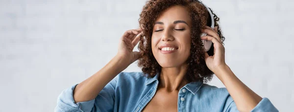 Кучерява афроамериканська жінка посміхається під час прослуховування музики в бездротових навушниках, банер — стокове фото