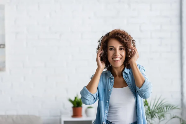 Mujer afroamericana positiva sonriendo mientras escucha música en auriculares inalámbricos - foto de stock