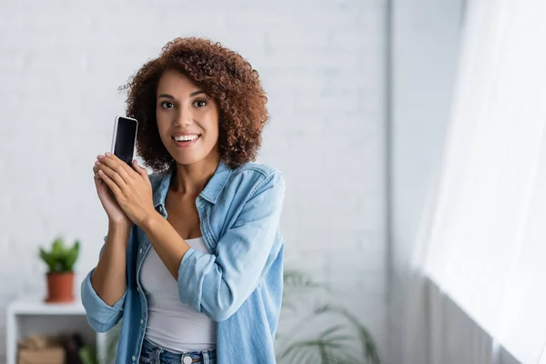 Mujer afroamericana emocionada sosteniendo teléfono inteligente con pantalla en blanco y mirando a la cámara - foto de stock