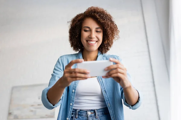 Visão de baixo ângulo da mulher americana africana feliz com cabelo encaracolado segurando smartphone durante o bate-papo por vídeo — Fotografia de Stock