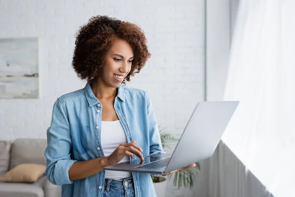 Heureuse femme afro-américaine avec les cheveux bouclés tenant ordinateur portable tout en travaillant à la maison — Photo de stock