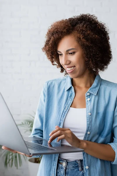 Fröhliche afrikanisch-amerikanische Frau mit lockigem Haar hält Laptop während der Arbeit von zu Hause aus — Stockfoto