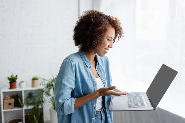 Seitenansicht einer lächelnden Afroamerikanerin mit lockigem Haar, die einen Laptop hält, während sie von zu Hause aus arbeitet — Stockfoto