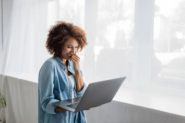 Donna afro-americana sorridente con i capelli ricci che tengono il computer portatile mentre lavora da casa — Foto stock