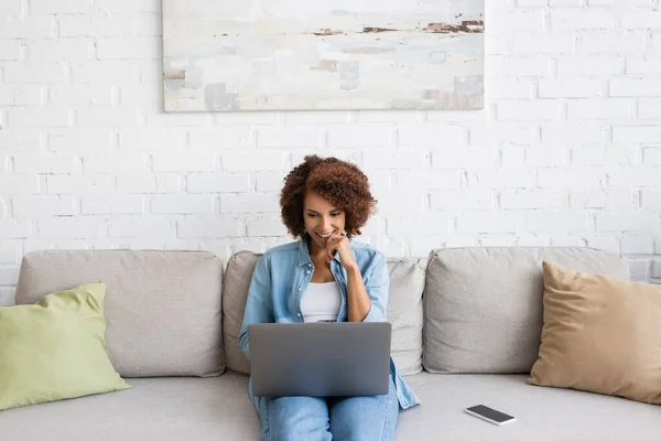 Heureuse femme afro-américaine en utilisant un ordinateur portable tout en étant assis sur le canapé et en travaillant à la maison — Photo de stock