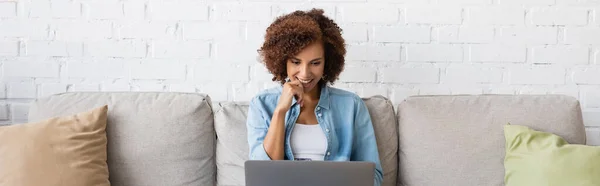 Happy african american woman using laptop while sitting on couch and working from home, banner — Stock Photo