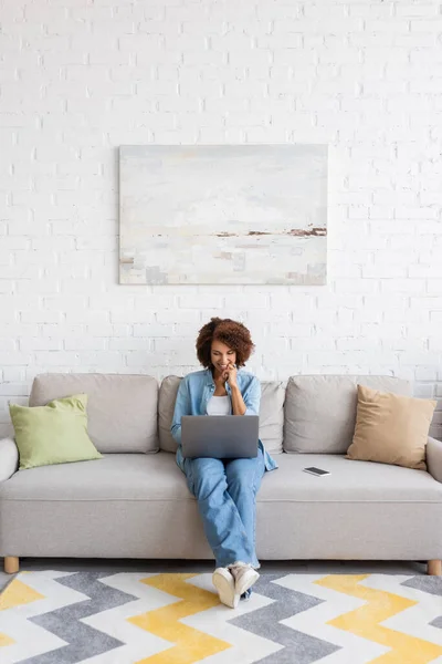 Весела афроамериканська жінка, яка користується ноутбуком, сидячи на дивані і працюючи вдома. — стокове фото