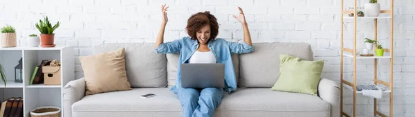 Excitée femme afro-américaine en utilisant un ordinateur portable tout en étant assis sur le canapé et en travaillant à la maison, bannière — Photo de stock