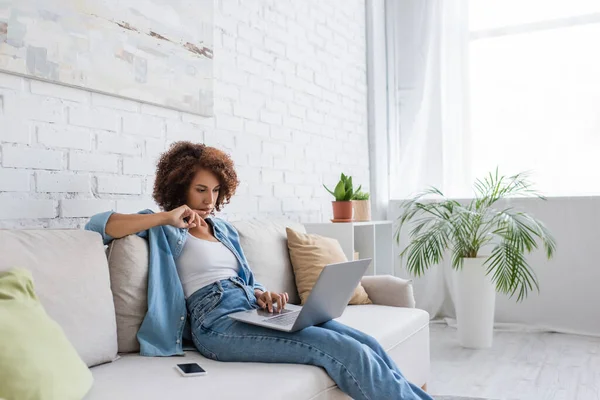 Pensive african american woman using laptop while sitting on couch and working from home — Stock Photo
