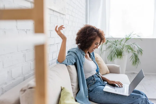 Side view of cheerful african american woman using laptop while sitting on sofa and working from home — Stock Photo